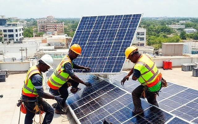 Professional photo: Ghanaian electricians installing solar panels on a commercial rooftop, teamwork and safety helmets, organized cable reels and toolboxes visible to suggest procurement/logistics efficiency, bright natural daylight, crisp detail, modern commercial setting, 16:9 composition